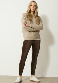 Beige oversized trui met contrasterende witte bies, gecombineerd met bruine nepleer legging. Model draagt witte sneakers met bruine zolen.