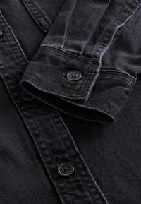 Gros plan sur la manche d'une chemise en denim noir avec un poignet noir boutonné et trois boutons noirs visibles sur la patte de boutonnage avant.