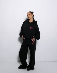 Czarny zestaw z bluzą i spodniami dresowymi o luźnym kroju. Bluza ma czerwone logo na piersi. Miękki, teksturowany materiał; casualowy design.