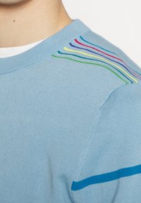 Épaule de pull en maille bleu clair avec de fines rayures multicolores près du col et une seule rayure bleue plus foncée autour du haut du bras.