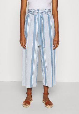 Frau trägt hoch taillierte, weit geschnittene, kurze Hose mit blau-weißen vertikalen Streifen, gebundenem Gürtel und braunen Sandalen mit Knöchelriemen.