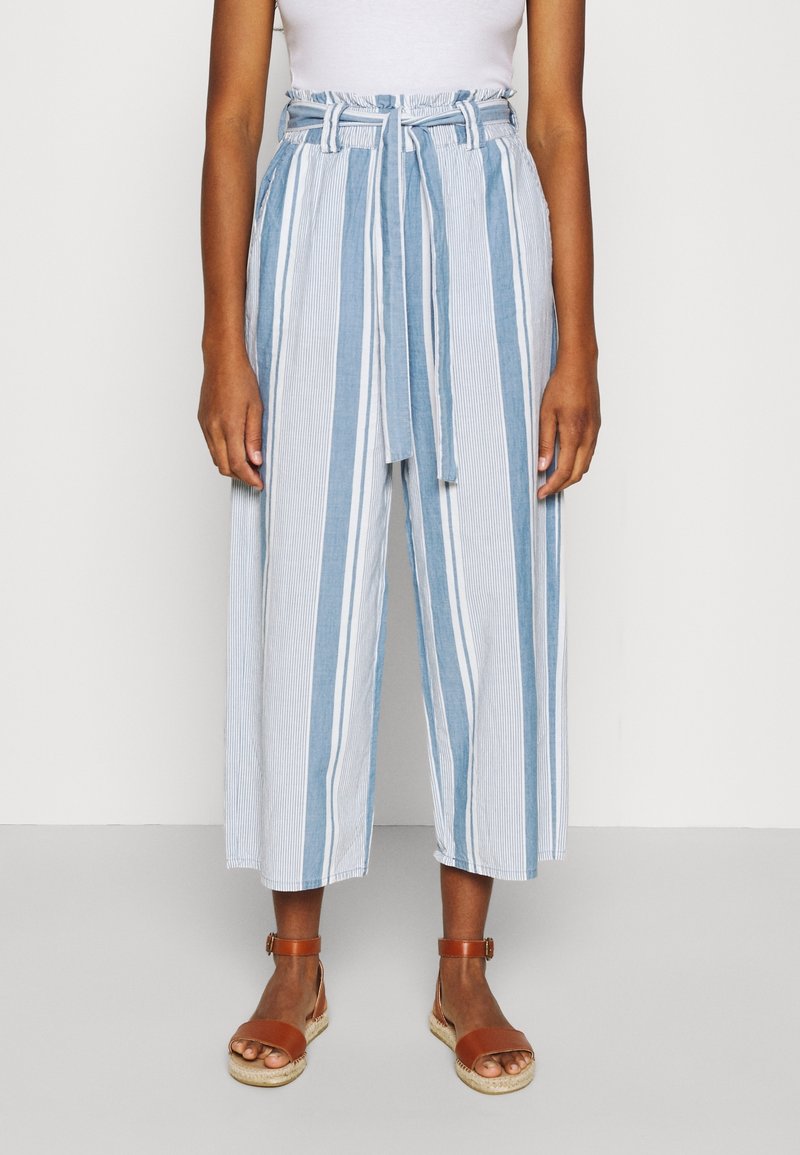 Femme portant un pantalon court taille haute à jambes larges avec des rayures verticales bleu et blanc, ceinture nouée, sandales marron à bride cheville.