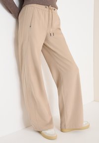 Beige široké kalhoty s �šňůrkou v pase, nošené s bílými teniskami a hnědým dlouhým rukávem, model opřený o bílou zeď.