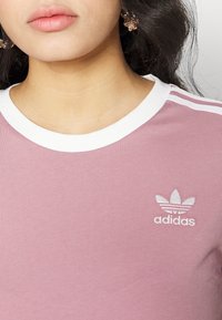 Rožinė medvilninė marškinė su apvaliu baltu apykakle, ant kairiojo šono turinčios balta siuvinėtą Adidas logotipą ir baltas pečių juosteles.
