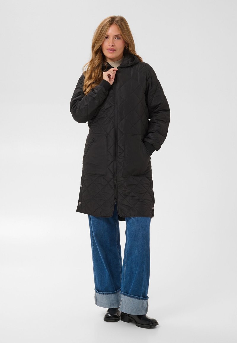Une femme porte un long manteau noir matelassé à capuche, un jean à jambes larges avec les revers retroussés et des chaussures noires, sur un fond clair uni.