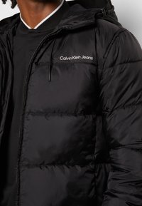 Giacca imbottita nera con texture trapuntata, dotata di cappuccio e chiusura con zip. Logo "Calvin Klein Jeans" bianco sul lato sinistro del petto.