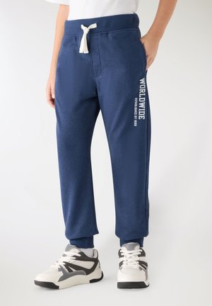 Bambino che indossa pantaloni della tuta blu navy con cordoncino bianco e testo "WORLDWIDE" sulla gamba, abbinati a scarpe da ginnastica bianche e grigie.