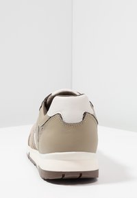 Beżowy but sportowy z teksturowaną siateczką na wierzchu, brązową podeszwą i białymi akcentami. Charakteryzuje się zaokrąglonym obcasem i kontrastującymi detalami przeszyć.