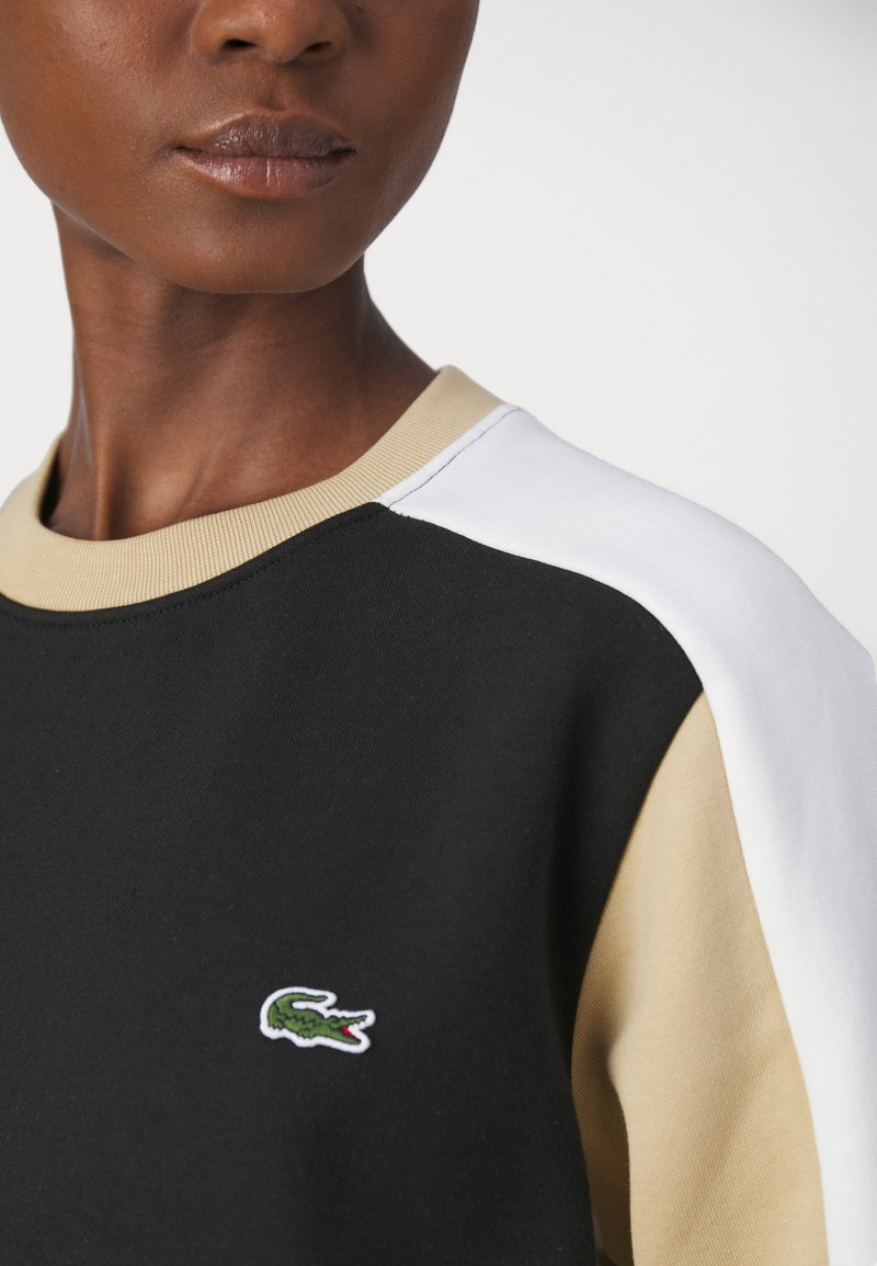 Sweat-shirt noir avec des accents beiges et blancs, encolure ronde et petit logo de crocodile vert sur la poitrine. Texture de tissu lisse.