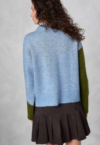 Pull en maille bleu avec des manches vertes, coupe courte et ourlet côtelé. Associé à une jupe plissée marron foncé. Tissu texturé.