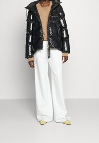 Osoba v lesklé černé péřové bundě, béžovém svetru, širokých bílých kalhotách a špičatých světlých žlutých botách na jednobarevném pozadí.