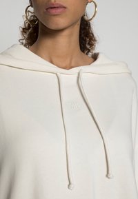 Sudadera con capucha color crema hecha de suave algodón, con capucha ajustable con cordón, detalle de logo en el frente y acentos acanalados alrededor del escote.