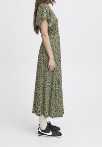 Grön blommig maxiklänning med korta svängda ärmar, midja med dragsystem och en svängande kjol, kombinerad med svarta sneakers och vita ankelsockor.