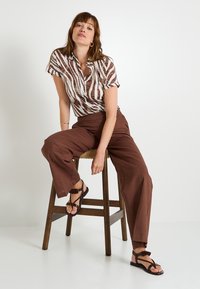 Camisa de manga corta a rayas marrones, pantalones de tiro alto marrones y sandalias negras con detalle de borlas. Taburete de madera y fondo neutro.