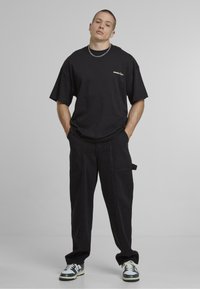 Tricou negru supradimensionat cu logo brodat îndrăzneț, asortat cu pantaloni cargo negri cu buzunare și o curea laterală, purtat cu adidași multicolori.