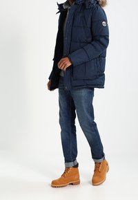 Doudoune bleu marine avec capuche bordée de fourrure, associée à un jean bleu et des bottes couleur fauve. La veste présente un design matelassé et un logo sur la manche.