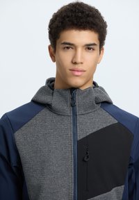 Giovane uomo con capelli ricci che indossa una giacca grigia e blu navy con zip e colletto alto su uno sfondo chiaro e semplice.