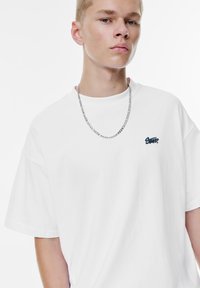 Hvid oversized T-shirt med korte ærmer, der har et lille blåt broderet logo på brystet. Komplementeret af en sølv kæde halskæde.
