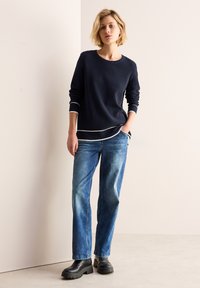 Marinblå ribbad tröja med vit kant, kombinerad med ljusblå raka jeans och svarta kängor med tjock sula. Mjuk textur, avslappnad design.