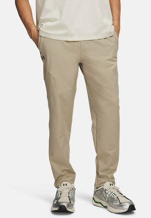 Man draagt beige Under Armour broek en witte, zilveren en groene hardloopschoenen, staat met handen in de zakken.