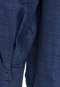 Κλείσιμο ενός σκούρου μπλε υφάσματος με ορατή υφή και δύο μαύρα κουμπιά σε μανσέτα μανικιού.