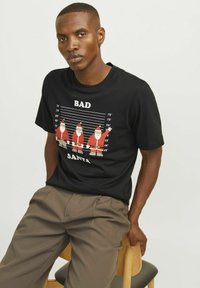 Sort bomulds T-shirt med en grafik af fire jules figure i rød med en linje-baggrund og teksten "BAD SANTA" nedenfor i hvid.