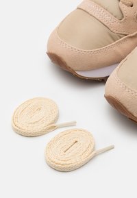 Beige treningesko har en teksturert semsket overdel og en glatt stoffoverdel. Inkluderer to rullede beige skolisser med plastspisser.