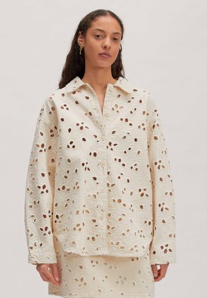 Femme portant une chemise crème boutonnée et une jupe assortie avec des motifs floraux en découpes ajourées, debout sur un fond uni.