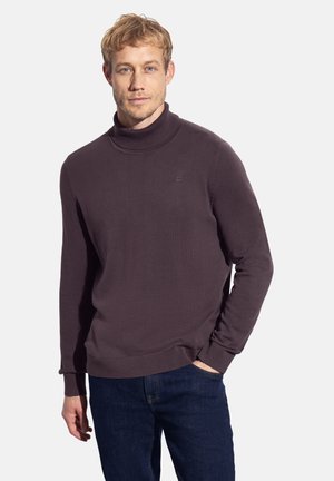 Suéter de cuello alto en un color púrpura oscuro, confeccionado con tejido suave, que presenta un cuello y puños acanalados, con un pequeño logo en el pecho. Combinado con unos jeans azules.