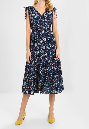 Femme portant une robe mi-longue à fleurs bleu marine avec taille froncée et liens aux épaules, associée à des chaussures à talons hauts jaunes sur fond blanc.