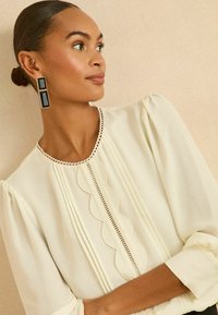 Bluză ivoire cu mâneci bufante, detalii texturate în formă de valuri și un dantelă delicată. O pereche de cercei dreptunghiulari negri cu accente aurii.