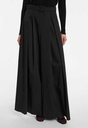 Femme portant un pantalon plissé noir à jambes larges avec une ceinture assortie, associé à des chaussures foncées et un haut noir à manches longues, debout sur fond blanc.