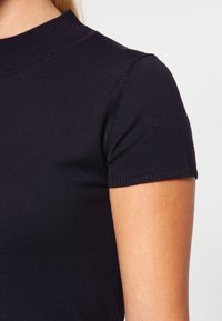 Mørkeblå strikket bluse med høj hals og korte ærmer samt ribbet kant. Glat tekstur og tætsiddende design, der er velegnet til lag-på-lag.