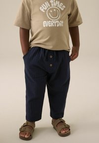 Beige T-Shirt mit Smiley-Grafik, marinefarbene, strukturierte Hose mit Kordelzug und Holzknöpfen sowie braunen Sandalen mit doppelten Schnallen.