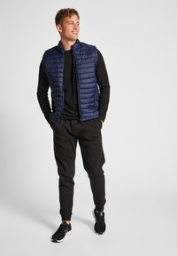 Marinblå quiltad väst med hög krage över en svart långärmad tröja, kombinerad med svarta joggers och svarta sneakers.