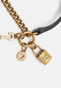 Kulta ketjuvaruste, jossa on lukko merkitty "EST 1981", kysymysmerkki-riipus ja kolmion muotoinen toistonappi, musta nahka-akcentti.