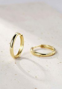Orecchini a cerchio in oro con una finitura liscia e lucida. Il design presenta una forma arrotondata e una chiusura sicura, appoggiati su una superficie tessurizzata.