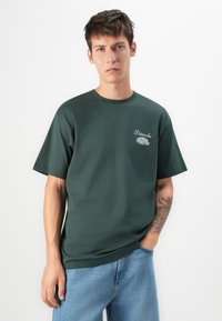 Young man with short dark hair wearing dark green "Diavola" T-shirt and light blue jeans, standing with one hand in pocket against white background.
