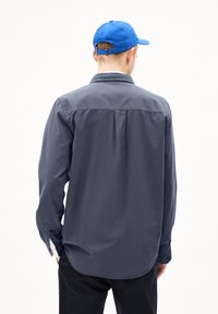 Donkergrijze langemouwenshirt met een button-down kraag, rugplooien en details op de manchetten. Draagt een blauwe pet met een verstelbare band.