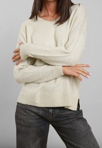 Maglione in maglia beige con scollatura rotonda, maniche lunghe e dettagli luccicanti. Presenta orli a coste e spacchi laterali. Indossato con jeans scuri.