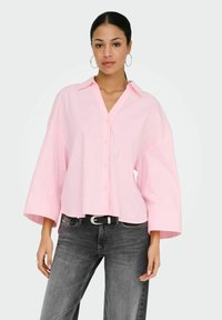 Femme portant une chemise boutonnée oversize rose pâle à manches larges et un jean noir délavé, debout devant un fond clair uni.