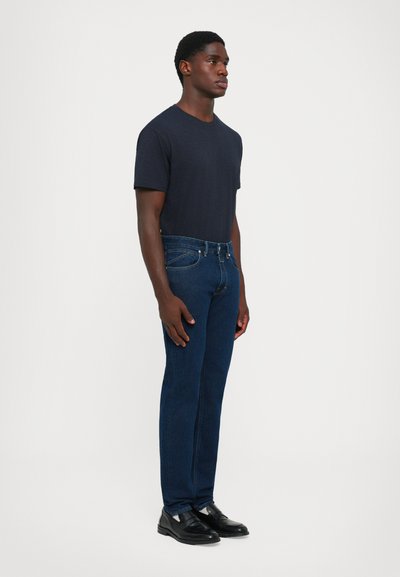 Navy crew-neck t-shirt, blue denim jeans, black dress shoes. Jeans feature traditional five-pocket design and subtle stitching details.