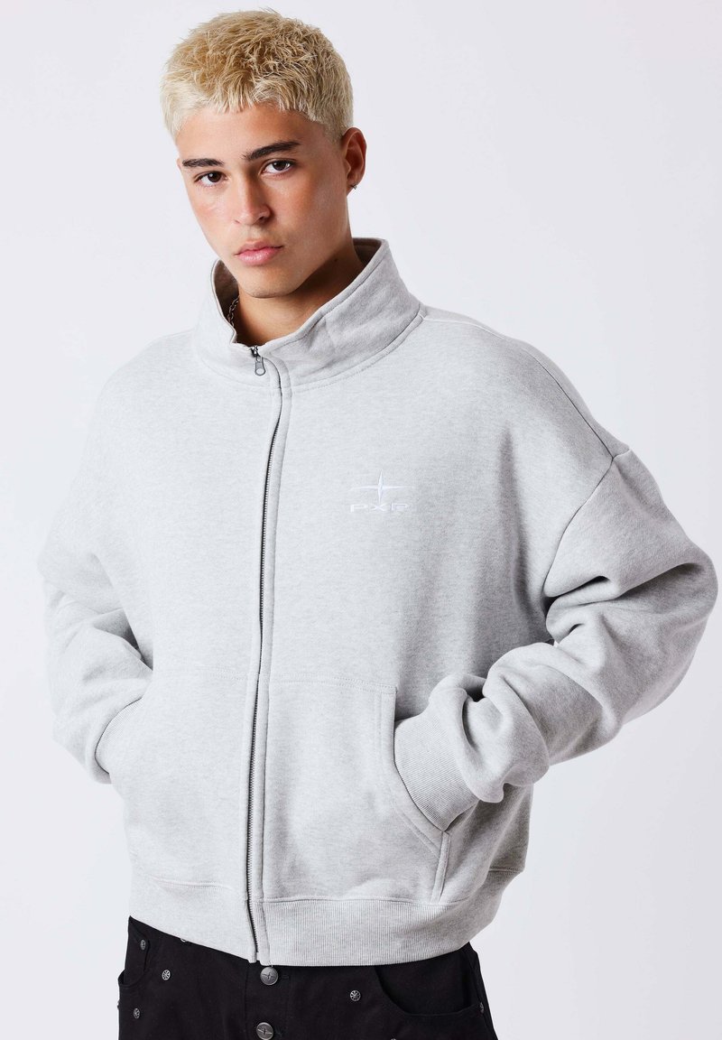 Jeune personne aux cheveux blonds courts portant un sweat-shirt gris clair zippé, les mains dans les poches, sur un fond uni.