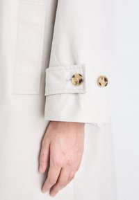 Kerge beeži värvi trench coat'i varrukas, mille mansettis on kaks suurt läikivat kilpkonnamustrit nuppu ja tekstuurne kangas.