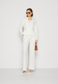 Weißes maßgeschneidertes Blazer- und Hosen-Set, ergänzt durch ein leichtes Tanktop. Kombiniert mit hohen weißen Schuhen und einer braunen Handtasche.
