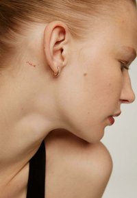 Aro de oro con pequeñas piedras de imitación, colocado junto a un tatuaje rojo tenue sobre piel clara, mostrando una textura suave y un diseño sencillo.