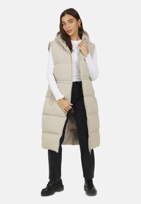 Panca lunga imbottita beige con cappuccio, caratterizzata da una chiusura con zip e tasche laterali. Indossata sopra un top bianco a maniche lunghe e pantaloni neri.