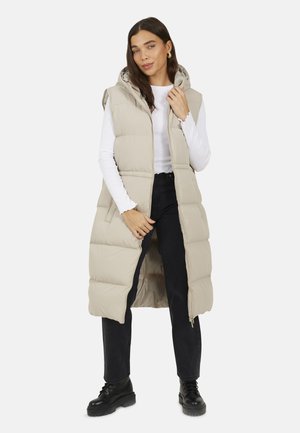 Beige gepolsterte lange Weste mit Kapuze, ausgestattet mit einem Reißverschluss und seitlichen Taschen. Getragen über einem weißen Langarmoberteil und schwarzen Hosen.