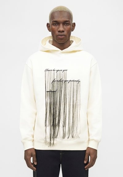 Moški s kratkimi blond lasmi, ki nosi belo kapuco s črnimi navpičnimi resicami in napisom: „Mir naj bo s tabo za tisto, kar si potrpežljivo prestal.“