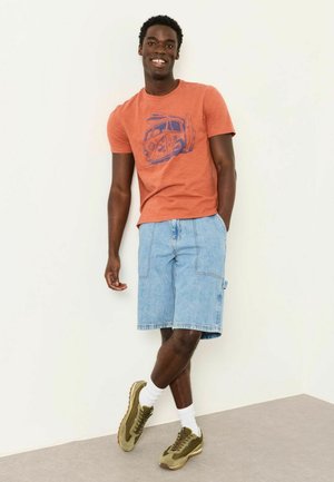 Junger Mann lächelnd, trägt orangefarbenes T-Shirt mit Grafik, hellblaue Jeansshorts, weiße Socken und olivgrüne Turnschuhe, steht vor einer weißen Wand.